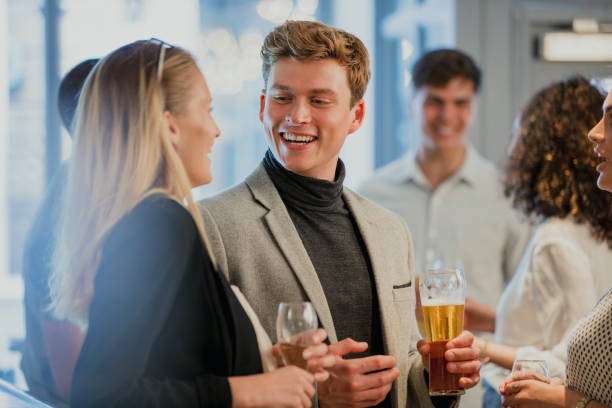 Coworkers are enjoying drinks in a bar after work. A man is holding a pint of beer and is talking to a woman who has her back to the camera.